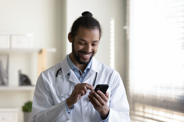Obraz premium Smiling young African American male general practitioner doctor physician using cellphone, reading email, giving professional healthcare consultation to client, web surfing, telemedicine concept.