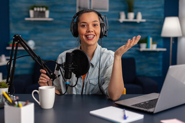 Portrait of woman smiling at camera in home recording studio. Vlogger with headphones creating content for online social media. Influencer streaming podcast using microphone and laptop.