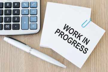 a white card with the text WORK IN PROGRESS on a wooden table next to a pen and calculator