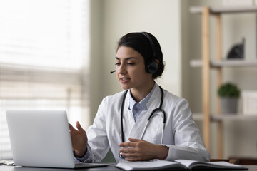 Happy young female Indian family doctor therapist in headphones with mic consulting patient online...