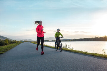 Happy mother and son go in sports outdoors. Boy rides bike in helmets, mom runs on sunny day. Silhouette family at sunset. Fresh air. Health care, authenticity, sense of balance and calmness
