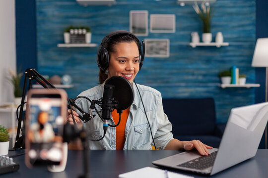 Woman vlogger with headphones looking at laptop and recording video. Influencer streaming online podcast on smartphone. Content creator in home studio using audio broadcasting equipment.