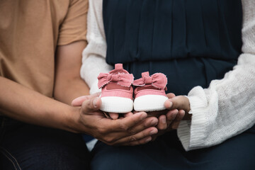 Close up, Pregnant women sit on bed with handsome husband and hold couple red baby shoes on the belly. A gift for the baby to be born in the future.