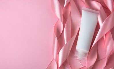 Cosmetic cream tube packaging with silk ribbon on pastel pink background. Beauty, skincare and...
