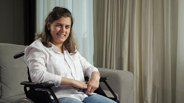Young disabled woman with infantile cerebral paralysis in white shirt smiles sitting in wheelchair near stylish sofa in living room at home.