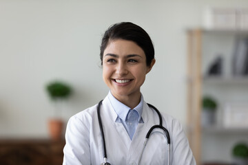 Head shot happy young female Indian GP doctor therapist in white medical coat looking in distance, thinking of career opportunities or professional challenges, feeling confident at workplace.