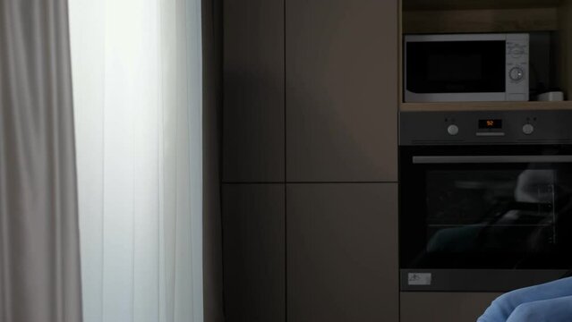Young disabled woman with cerebral palsy moves in wheelchair closer to window behind white curtain against kitchen furniture at home.