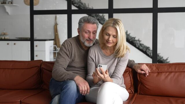 Hilarious mature couple sitting on the sofa and holding smartphone, smiling grey-haired man and cheerful blonde woman making video call by phone, middle-aged spouses watching pictures together
