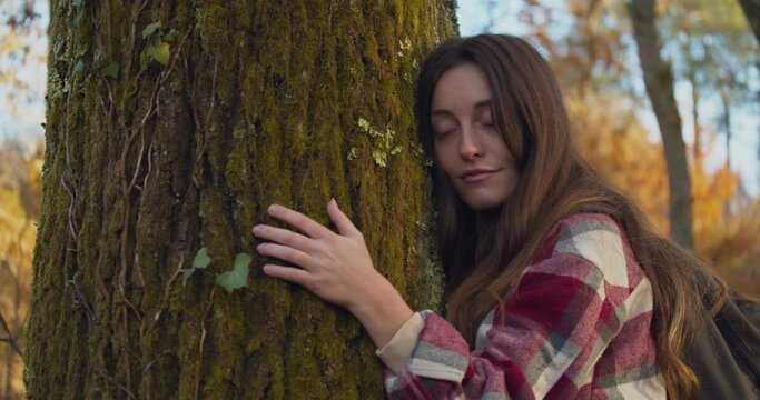 A young female with long hair hugs a tree and closes her eyes, medium shot
