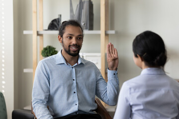 Happy millennial handsome African American man with hearing disability making gestures, using sign language practicing communication with professional female Indian therapist in clinic office. - Powered by Adobe