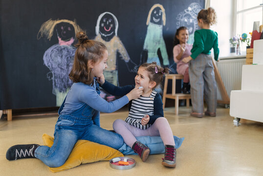 Group of little girls drawing with chalks on each other's cheeks indoors in playroom.