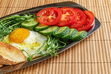 fried egg, toasted bread, cucumber and tomato slices and microgreens on a metal plate on bamboo straw surface, healthy breakfast concept