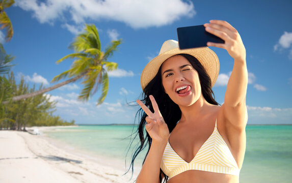travel, tourism and summer vacation concept - happy smiling young woman in bikini swimsuit and straw hat taking selfie with smartphone over tropical beach background in french polynesia