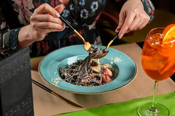 Cropped shot of woman eating black pasta with seafood served with orange spritz