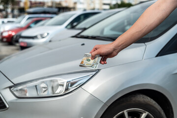 male hand holding a lot of dollars against gray car in the back