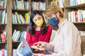 young couple of multiracial students using laptop in a library during Corona Virus epidemic, wearing face mask fro protection, team work and health care concepts - Powered by Adobe