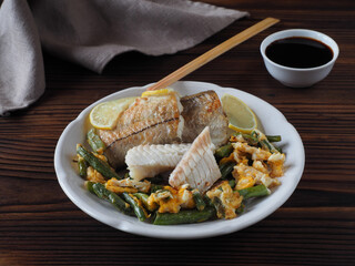Pieces of fried haddock fish (melanogrammus aeglefinus), green string beans with egg in a white plate, soy sauce, chopsticks on a dark wooden table, flat layout. Diet dish with vegetables and seafood