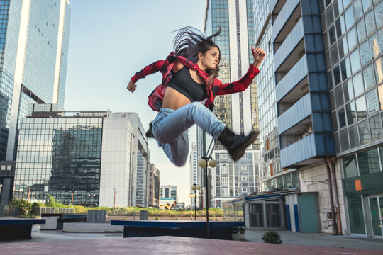 Photo shoot of stylish caucasian hip hop dancer posing jumping with red shirt, jeans and black boots shoes and top with a urban town as background. City shooting of moving model