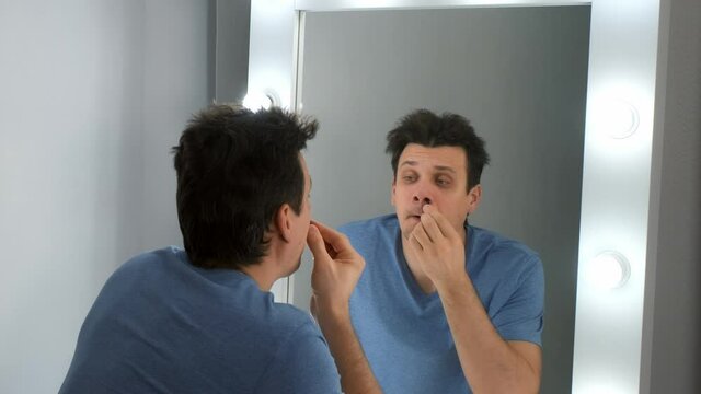 Man plucks the hair out of his nose using tweezers looking at mirror with light bulbs at home. The concept of male self-care and hygiene. He is in pain, but continues his actions.