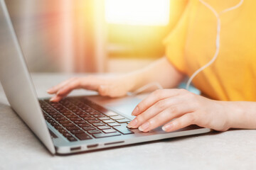 A woman is sitting at a desk with headphones on, typing on a laptop. A window in the background. Hands close-up. Copy space. The concept of online education