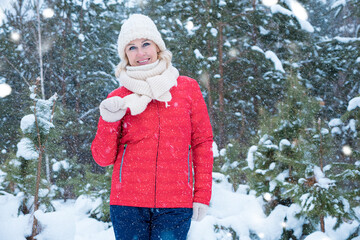 Cheerful woman in knitted hat and wrapped with scarf in mittens stands against coniferous trees...