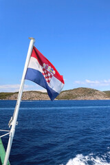 Fototapeta premium Flag of Croatia blowing in the wind. Adriatic sea, island and boats in the background.