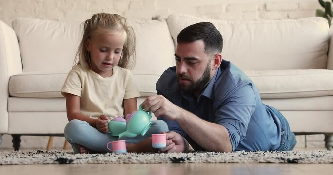Preschool daughter sitting on floor carpet play tea ceremony game with loving young father holds teacup pour imaginary tea, play game in living room, looking playful, enjoy time together. Fun concept