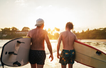 Father with teenager son walking with surfboards by the sandy ocean beach with palm trees on background lightened with sunset sun. They smiling and have a conversation. Family active vacation concept.