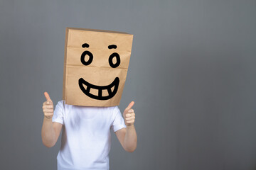 A boy in a white T-shirt with a paper bag on his head with a painted face and a smile shows a like with his thumbs