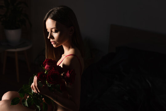 Seductive Woman Sitting In Dark Bedroom With Bouquet Of Red Roses.