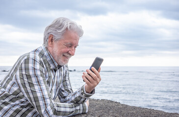 Adult mature pensioner senior man smiling enjoying outdoors at sea using mobile phone. Active bearded grandfather relaxing in retirement wearing a checkered shirt. Cloudy sky and horizon over water