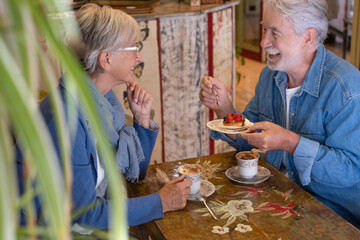 Adult senior couple in coffee shop having breakfast together  drinking coffee and cappuccino. Two attractive smiling elderly people enjoying retirement and stay together. Fruit cake on man hands