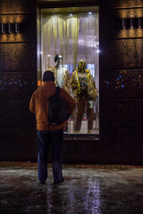 The figure of a man in an old orange jacket and with a backpack in front of a brightly lit window of an expensive fashion store on a dark background