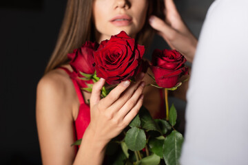 cropped view of blurred woman holding red roses near man on dark background.