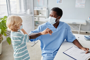 Portrait of young African-American doctor bumping elbows with cute little kid as contactless greeting during visit to pediatrician in medical clinic