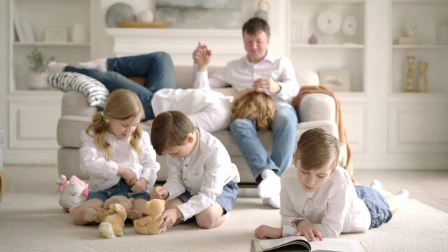Caucasian cute happy family got together at home. Mother and father sit on sofa hugging watching their young children, girl and two boys playing with soft toys and reading book at home in living room