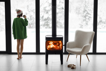 Woman standing with cup by the fireplace near the window at modern house on nature during winter...