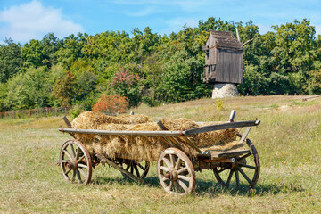 Fototapeta na wymiar An old wooden cart loaded with straw against the background of a summer landscape with an old wooden mill.