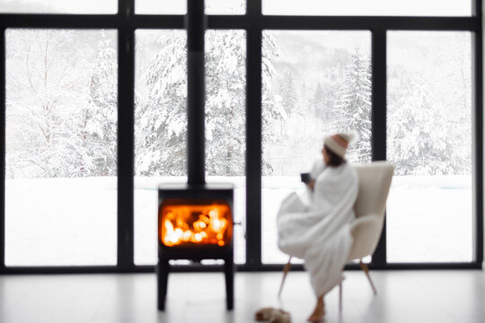 Woman covered with blanket enjoys winter time at home with burning fireplace, looking outside the window on snowy landscape. Concept of winter mood. Idea of winter vacation in the mountains