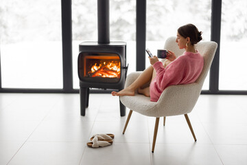 Woman sitting with cup on chair by the fireplace at modern house on nature during winter time....