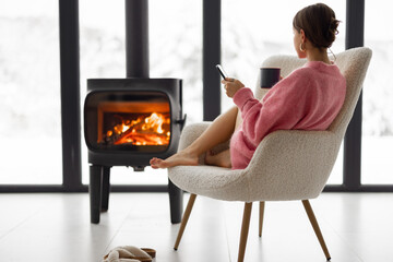 Woman sitting with cup on chair by the fireplace at modern house on nature during winter time....