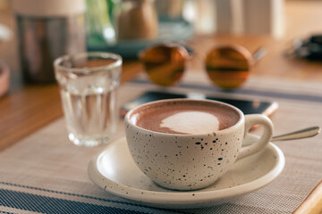 A cup of coffee on a wooden table of a street cafe on a summer day. A glass of water, sunglasses and smartphone in the background