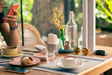 Good morning at the cafe in summer. A cup of coffee on a wooden table. Woman's hand with smartphone, a bottle of water and glass, sunglasses and smartphone in the background