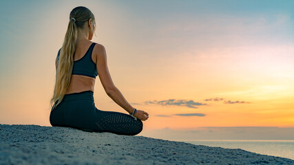 Fototapeta na wymiar Young caucasian woman with long hair meditating in a black clothes for yoga on a rocks on the top by the sea. On the background colorful sunrise sky.