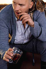 A young man smokes a cigar and holds a glass of alcohol. A cat is sitting on his shoulder.