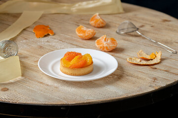 A tartlet with tangerine and grapefruit lies on a plate on a wooden table, around the tools and ingredients for making a dessert