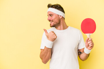 Young caucasian man holding ping pong racket isolated on yellow background points with thumb finger...