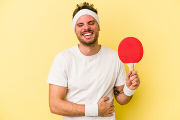 Young caucasian man holding ping pong racket isolated on yellow background laughing and having fun.