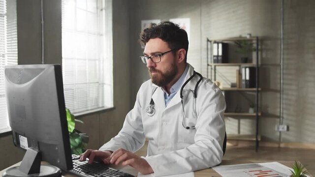 Smiling young man professional medic doctor wear white medical uniform glasses looking at camera siting in hospital office