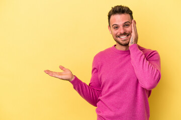 Fototapeta premium Young caucasian man isolated on yellow background holds copy space on a palm, keep hand over cheek. Amazed and delighted.